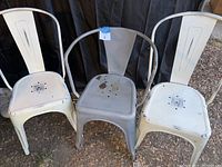 Three metal cafe chairs in a row: two white and one grey in the center, showing rust.