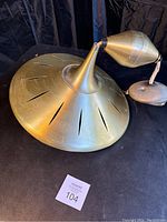 Full view of brass mid century ceiling light fixture on dark backdrop, showing saucer shade with cut-out slits and adjustable arm with conical joint.