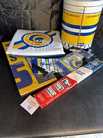 Photo of vintage LA Rams items including a blue and yellow seat pad, popcorn holder with yellow and blue stripes and 'Los Angeles' text, foldable lunch bags, and a boxed retractable car sun shade.