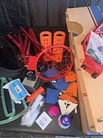 View of assembled sports equipment including cornhole bean bags, basketball hoops, orange glove drying sleeves, and various fitness accessories placed on and around the cornhole board.