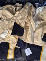 Two cream-colored UCLA band uniform jackets and matching navy blue pants with yellow stripe. One jacket stained with felt residue from covered UCLA embroidery, other with embroidery removed.