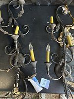 Two vintage wrought iron wall sconces with three yellow candle-style bulb holders each and some bulbs included, showing leaf and curled iron decorative elements on a dark background.