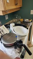 Photo showing three white mixing bowls stacked, metal roasting pans, manual can opener, and some kitchen items in a kitchen corner with wood cabinets and green wall
