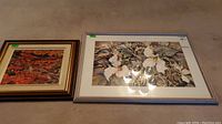 Two framed artworks on carpet floor. Left artwork is an abstract watercolor painting in warm colors, right artwork is a floral print in silver frame.