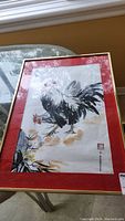 Framed watercolor artwork of a rooster on a table, showing full front view with red mat and gold frame.