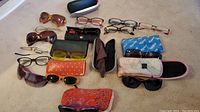 Several pairs of eyeglasses and sunglasses on beige carpet, many with cases in various colors and materials.