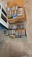 Photo showing stacked and boxed CDs along with boxes of cassette tapes on carpeted floor.