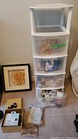 White plastic storage bin with four drawers filled with various sewing accessories, beside a yellow flower shadow box frame and small boxes with sewing tools