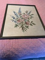 Front view of antique needlepoint floral embroidery artwork in a dark wooden frame placed on pink quilted fabric.