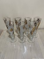 Set of seven tall clear pilsner glasses decorated with gold leaf motifs around the bowl and gold rims.
