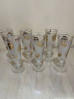 Front view of six tall clear pilsner glasses with gold leaf and gold rim decoration, arranged in single row.