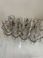 Eight clear glass tumblers with gold leaf design in brass holder, showing overall condition and arrangement.