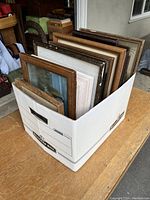 Box filled with 20 small framed art pieces, arranged vertically inside a white storage box on wooden table.