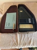 Top view of Oscar Schmidt autoharp inside open brown hard case with instruction manual, tuning key, and guitar pick.