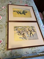 Two framed prints laid flat on a floral quilted surface, showing part of each artwork and frame.