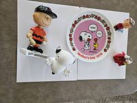 Top-down view of Peanuts collectibles including Charlie Brown figurine, Snoopy eyeglass holder, 1978 Mother's Day plate, Linus figurine, and Charlie Brown bobblehead.
