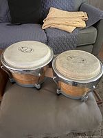 Front angle view of pair of percussion bongos with natural skins and wooden shells clustered together on cushioned surface.