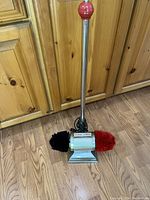 Full view of the vintage Dremel electric shoe shine buffer standing on wooden floor against a wooden cabinet, showing red knob on top and dual brushes at base.