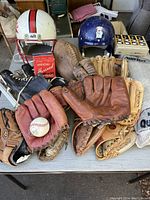 Wide shot showing white football helmet, blue baseball helmet, several baseball gloves, football, vintage baseball, sports net, and black ice skates