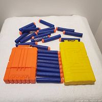 Group of 24 blue Nerf Elite foam darts with orange tips, scattered with two plastic Nerf clips—one orange clip holding six darts, one yellow clip holding eight darts—laid on a white surface.