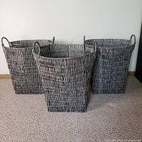 Three grey woven storage baskets made of synthetic material on metal frames, arranged on carpeted floor against white wall. One basket is square-shaped and two are round-shaped with handles on sides.