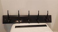 Glossy black 5-hook coat hanger mounted horizontally on black wooden base against white background, showing size in relation to ruler below.