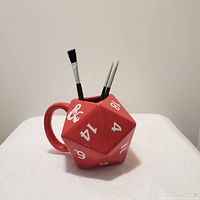 Red 20-sided die shaped ceramic mug with white numbers and D&D logo, with paintbrushes inside, on white surface against plain background.
