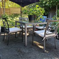 Patio dining set showing metal table with glass top and four chairs with white cushions outside in garden setting.