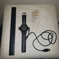 Garmin Fenix GPS hiking watch with a 10-inch black rubber adjustable band, digital liquid crystal display, accompanied by a charging cord and quick start instruction manual, all laid out alongside a black ruler for scale.