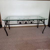 Photo shows a rectangular coffee table with a clear beveled tempered glass top and a charcoal powder coated metal frame incorporating scrollwork design on the sides and curved legs.