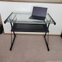 Modern black metal desk with clear tempered glass top and black metal mesh lower shelf. Laptop on top for scale. Carpet floor background.