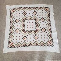 Full view of the square linen hand-embroidered tablecloth laid flat on carpet, showing embroidered geometric diamond and floral designs in red, green, gold, and black thread.