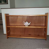 Photo of a long natural wood shelving unit with two shelves, single teapot placed on upper shelf for scale, showing front and side profile against a neutral interior background.