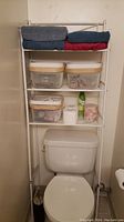 Full view of the metal bathroom storage unit fitted over the toilet, showing four shelves filled with towels, plastic baskets, and bathroom essentials.