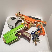 Top view of three Nerf N-Strike Elite toy blasters and gray belt holster on a white surface. Toys feature gold accent paint highlights. Shows various colors of the blasters including green, orange and blue along with beige main body parts.