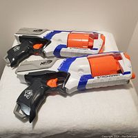 Two Nerf Strongarm blasters side by side on white fabric background, showing detailed view of orange six-dart cylinder and labeling.