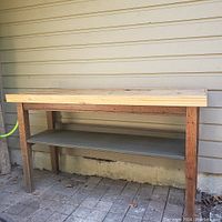 Side angle photo of vintage oak work table showing the rough stripped wooden top, oak legs, and lower shelf against a beige exterior wall.