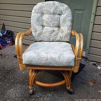 Front view of rattan armchair showing cushioned seat and back with leafy pattern fabric upholstery, curved armrests, and circular rattan base with wheels.