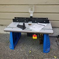 Front view of the Mastercraft router table showing adjustable guide fence pieces, safety guard, and manual on the table surface with blue legs.