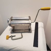 Metal clay roller with yellow crank handle, metal C-clamp, and black ruler placed on a white cloth surface.