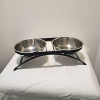 Photo of twin stainless steel pet feeding bowls set in black metal elevated stand on white cloth background.