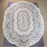 Ecru/beige oval acrylic lace tablecloth with intricate floral and geometric patterns placed over the mauve cotton tablecloth for contrast, measuring approximately 5' x 7'.