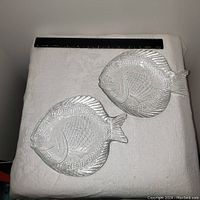 Two small clear embossed glass dishes shaped like fish shown on white fabric with a black ruler for scale.