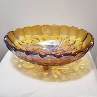 Front angle view of amber footed glass bowl with embossed fruit and leaves at bottom, showing scalloped rim with iridescent finish.