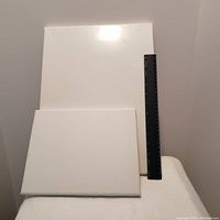 Photo of two white painting canvases, one 14x12 inches shrink wrapped and one smaller 10x8 inches, both blank and resting against a wall with a black ruler showing scale.