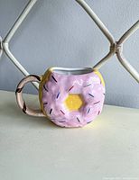 Front and side view of ceramic donut mug showing pink frosting and rounded chocolate glaze handle.