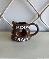 Front view of brown ceramic mug shaped like a poop emoji with 'HOLY CRAP!' text above and below a central hole.