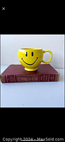 Side view of yellow smiley face mug showing front face design and single handle, sitting on a vintage book.