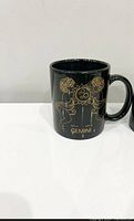 Black ceramic mug with Gemini zodiac sign and gold detailing, displayed on white surface