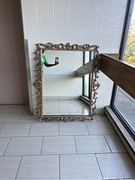 Full view of large rectangular ornate mirror leaning against a tiled wall near a window, showing overall condition and size.